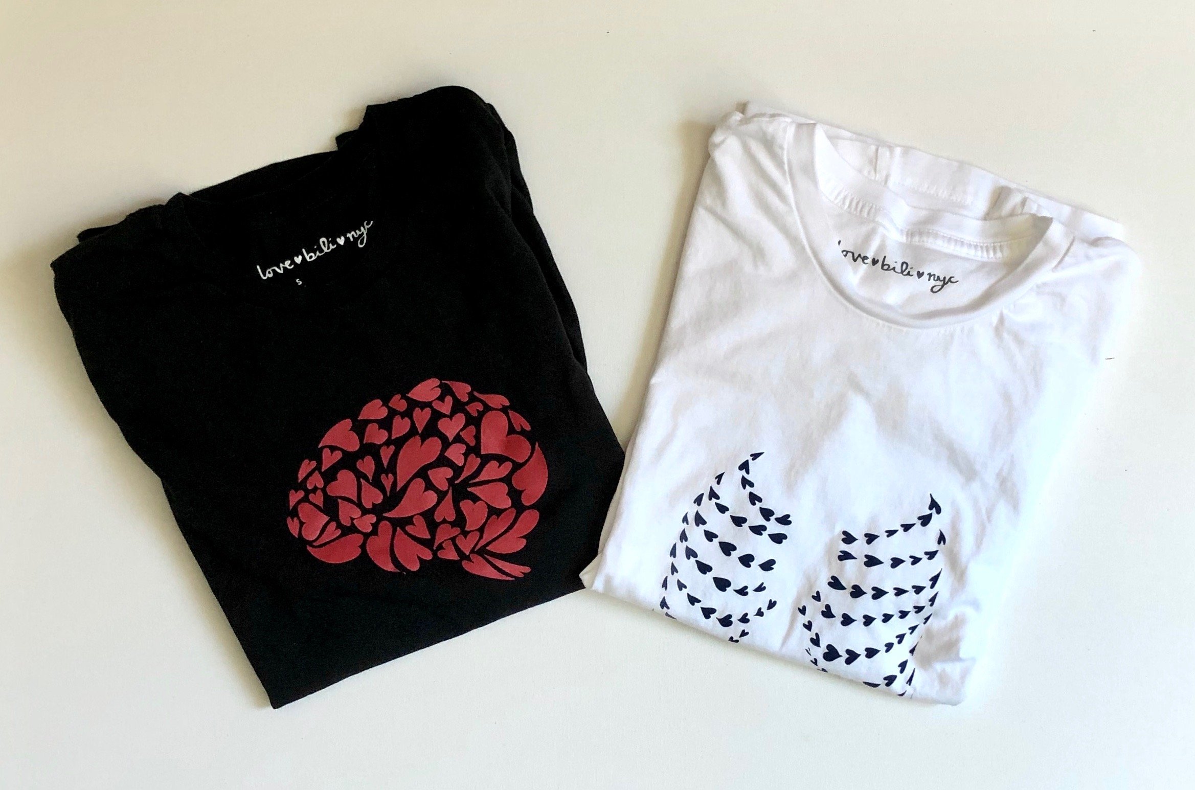 Two ♡bili shirts folded next to each other. The shirt on the left is black with a red brain design made of hearts. The shirt on the right is white with a black ribcage design made of hearts